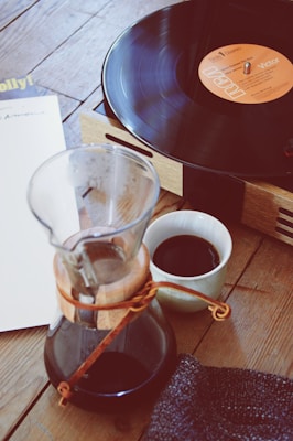 A wooden tabletop holds a record player with a vinyl playing. Nearby, there's a glass carafe with coffee and a small cup filled with coffee. A piece of fabric lies beside the cup. The scene suggests a cozy, relaxed setting.
