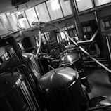 A black and white industrial setting features several large, shiny metal tanks connected by pipes and tubes. The room is spacious with large windows allowing in natural light. The tanks are likely used for processing or brewing, and various machinery components are visible around them. The reflective surfaces of the tanks provide a striking contrast in the monochrome image, adding an industrial aesthetic.