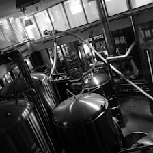 A black and white industrial setting features several large, shiny metal tanks connected by pipes and tubes. The room is spacious with large windows allowing in natural light. The tanks are likely used for processing or brewing, and various machinery components are visible around them. The reflective surfaces of the tanks provide a striking contrast in the monochrome image, adding an industrial aesthetic.