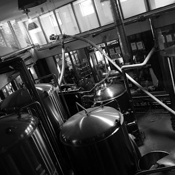 A black and white industrial setting features several large, shiny metal tanks connected by pipes and tubes. The room is spacious with large windows allowing in natural light. The tanks are likely used for processing or brewing, and various machinery components are visible around them. The reflective surfaces of the tanks provide a striking contrast in the monochrome image, adding an industrial aesthetic.