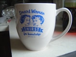 A white mug featuring blue illustrations of two women along with the text 'Smart Women Thirst for Knowledge' is on a kitchen countertop. The background includes a blurred view of various kitchen items.