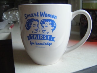A white mug featuring blue illustrations of two women along with the text 'Smart Women Thirst for Knowledge' is on a kitchen countertop. The background includes a blurred view of various kitchen items.