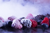 A variety of frozen berries including raspberries, blackberries, and blueberries are scattered on a surface. The berries are covered with a layer of frost, and mist surrounds them, creating a cold, wintry atmosphere.
