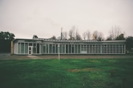 A vintage car dealership building with large glass windows and signage that reads 'Volkswagen Audi Kundendienst'. The structure is situated in a barren area with leafless trees in the background, under an overcast sky. The foreground features a patch of green grass.