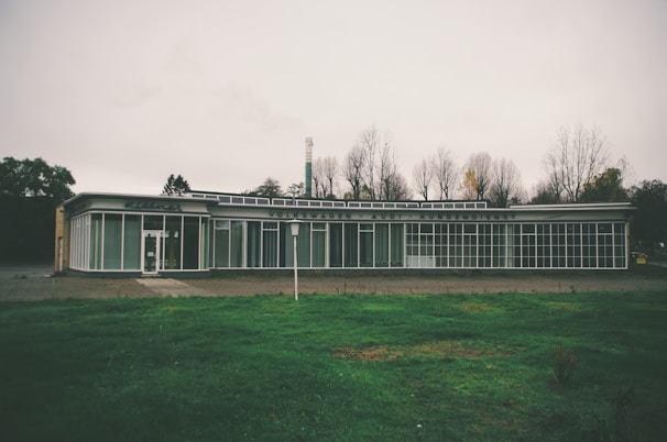 A vintage car dealership building with large glass windows and signage that reads 'Volkswagen Audi Kundendienst'. The structure is situated in a barren area with leafless trees in the background, under an overcast sky. The foreground features a patch of green grass.