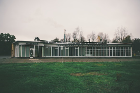 A vintage car dealership building with large glass windows and signage that reads 'Volkswagen Audi Kundendienst'. The structure is situated in a barren area with leafless trees in the background, under an overcast sky. The foreground features a patch of green grass.