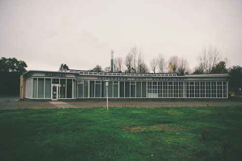 A vintage car dealership building with large glass windows and signage that reads 'Volkswagen Audi Kundendienst'. The structure is situated in a barren area with leafless trees in the background, under an overcast sky. The foreground features a patch of green grass.