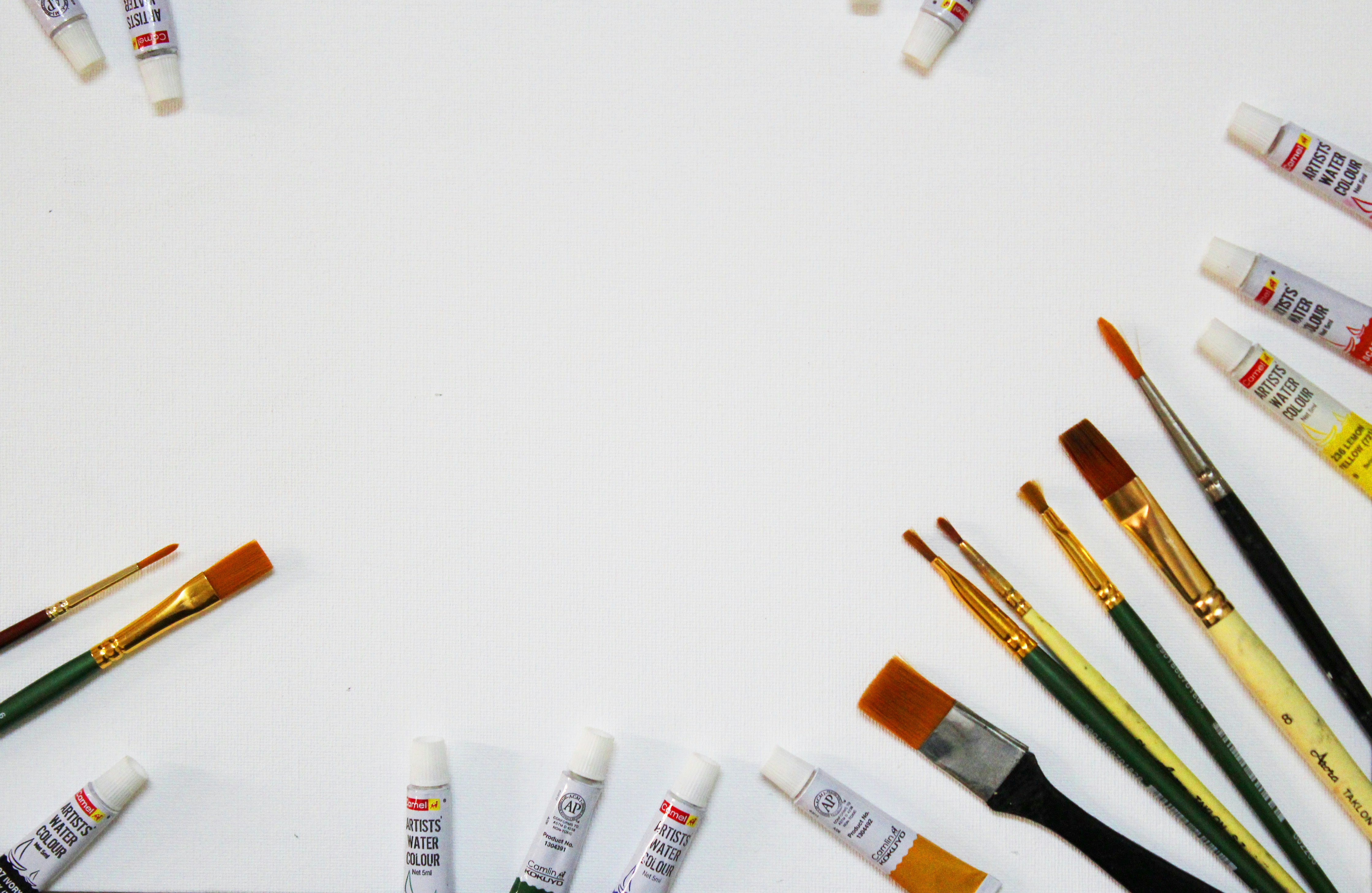 Various paintbrushes and tubes of artists' watercolor paints are scattered along the edges of a white surface, creating an arrangement that frames the central space.