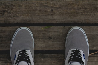 A pair of grey canvas shoes with white and black laces are positioned on a wooden surface with visible textures and grain patterns. A small green leaf is placed on the wooden surface between the shoes.