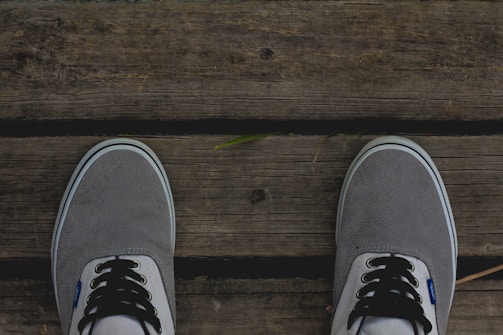 A pair of grey canvas shoes with white and black laces are positioned on a wooden surface with visible textures and grain patterns. A small green leaf is placed on the wooden surface between the shoes.