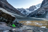 A serene mountainous landscape with snow-capped peaks in the background. A vibrant green tent is pitched on rocky terrain near a large boulder in the foreground, indicating a campsite. Sparse patches of snow and earth are visible, set against the imposing mountain scenery.