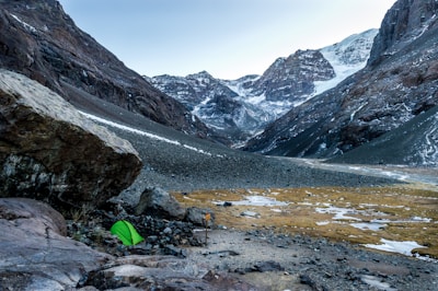A serene mountainous landscape with snow-capped peaks in the background. A vibrant green tent is pitched on rocky terrain near a large boulder in the foreground, indicating a campsite. Sparse patches of snow and earth are visible, set against the imposing mountain scenery.