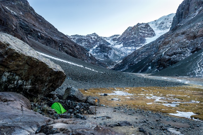 A serene mountainous landscape with snow-capped peaks in the background. A vibrant green tent is pitched on rocky terrain near a large boulder in the foreground, indicating a campsite. Sparse patches of snow and earth are visible, set against the imposing mountain scenery.