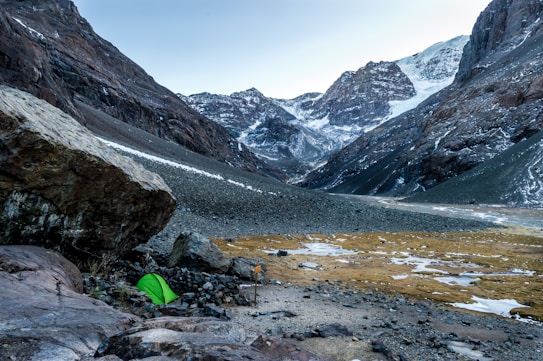 A serene mountainous landscape with snow-capped peaks in the background. A vibrant green tent is pitched on rocky terrain near a large boulder in the foreground, indicating a campsite. Sparse patches of snow and earth are visible, set against the imposing mountain scenery.