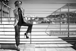 A fashionable woman stands on a rooftop, leaning against a metal railing. She wears sunglasses, a stylish hat, a layered outfit with a jacket and lace pants, and high heels. Her accessories include numerous bracelets on her arm. In the background, urban buildings and a clear sky can be seen.