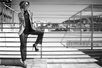 A fashionable woman stands on a rooftop, leaning against a metal railing. She wears sunglasses, a stylish hat, a layered outfit with a jacket and lace pants, and high heels. Her accessories include numerous bracelets on her arm. In the background, urban buildings and a clear sky can be seen.