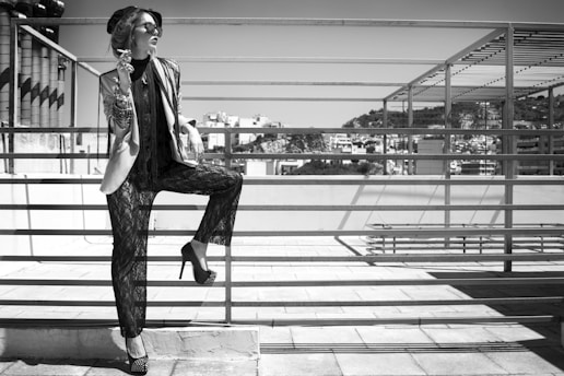 A fashionable woman stands on a rooftop, leaning against a metal railing. She wears sunglasses, a stylish hat, a layered outfit with a jacket and lace pants, and high heels. Her accessories include numerous bracelets on her arm. In the background, urban buildings and a clear sky can be seen.