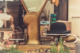 A vintage weighing scale is prominently featured with a bowler hat placed on the measuring platform. Rusty weights are stacked next to the scale, and the background shows a blurred view of a person working in a kitchen or butcher setting. The surroundings include metal equipment and some greenery, adding a rustic and nostalgic atmosphere.