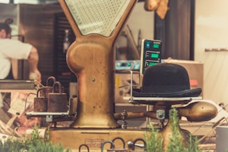 A vintage weighing scale is prominently featured with a bowler hat placed on the measuring platform. Rusty weights are stacked next to the scale, and the background shows a blurred view of a person working in a kitchen or butcher setting. The surroundings include metal equipment and some greenery, adding a rustic and nostalgic atmosphere.