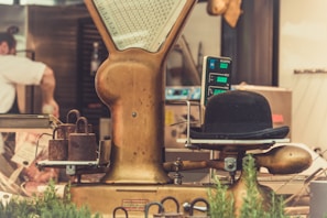 A vintage weighing scale is prominently featured with a bowler hat placed on the measuring platform. Rusty weights are stacked next to the scale, and the background shows a blurred view of a person working in a kitchen or butcher setting. The surroundings include metal equipment and some greenery, adding a rustic and nostalgic atmosphere.
