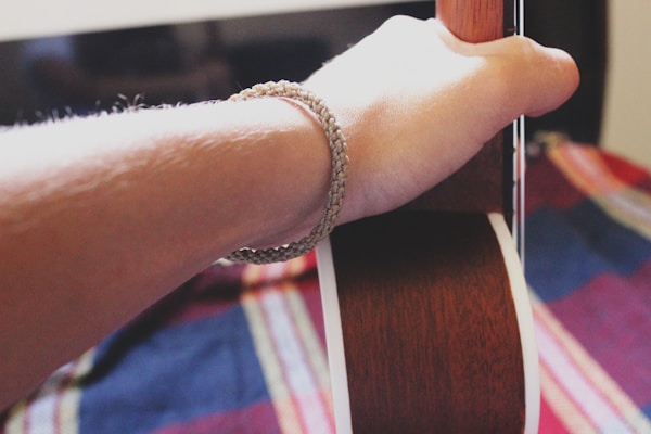 A forearm and hand are visible, holding the neck of a guitar. The person is wearing a woven bracelet. The background features a colorful, patterned fabric in shades of red, blue, and yellow.