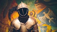 A medieval suit of armor stands prominently in the foreground, featuring a helmet with a visor and intricate metal plating. In the background, a colorful mural displays a knight in armor alongside elaborate heraldic designs, including a bird and various emblems.