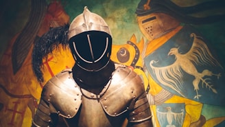 A medieval suit of armor stands prominently in the foreground, featuring a helmet with a visor and intricate metal plating. In the background, a colorful mural displays a knight in armor alongside elaborate heraldic designs, including a bird and various emblems.