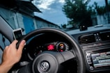 A hand grips the steering wheel of a car, with the dashboard in view showing a speedometer and a car radio screen displaying song information. A smartphone is mounted on the dashboard, and outside the car window, a road flanked by buildings is visible under a cloudy sky.