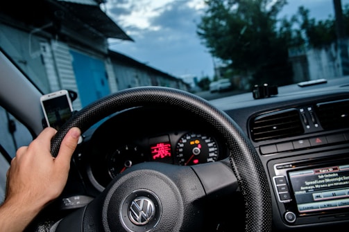 A hand grips the steering wheel of a car, with the dashboard in view showing a speedometer and a car radio screen displaying song information. A smartphone is mounted on the dashboard, and outside the car window, a road flanked by buildings is visible under a cloudy sky.