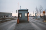 A small, metal security booth with a traffic barrier arm situated on a road leading to an industrial area. Surrounding the booth are road signs and industrial structures like cranes and warehouse buildings. The sky is overcast and trees without leaves are visible, indicating a wintry setting.