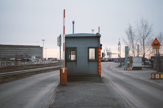A small, metal security booth with a traffic barrier arm situated on a road leading to an industrial area. Surrounding the booth are road signs and industrial structures like cranes and warehouse buildings. The sky is overcast and trees without leaves are visible, indicating a wintry setting.
