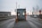 A small, metal security booth with a traffic barrier arm situated on a road leading to an industrial area. Surrounding the booth are road signs and industrial structures like cranes and warehouse buildings. The sky is overcast and trees without leaves are visible, indicating a wintry setting.