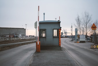 A small, metal security booth with a traffic barrier arm situated on a road leading to an industrial area. Surrounding the booth are road signs and industrial structures like cranes and warehouse buildings. The sky is overcast and trees without leaves are visible, indicating a wintry setting.