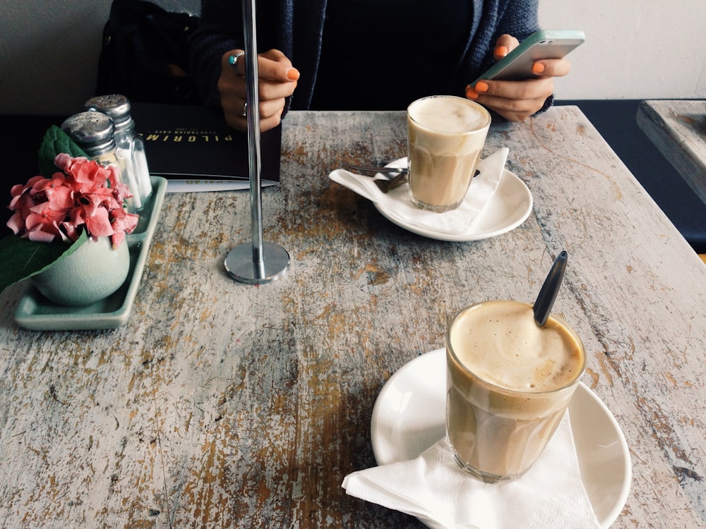 A person with a smartphone in their hand sitting at a table with two cups of latte