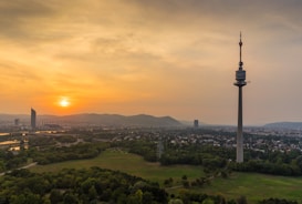 A serene landscape featuring a prominent telecommunications tower against a sunset. The sky is a mix of orange and gold hues, creating a tranquil atmosphere. Lush green fields and a mix of urban and natural scenery spread across the foreground, with distant mountains visible on the horizon.