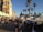 A busy outdoor marketplace with numerous people walking along a street lined with vendor stalls. Tall palm trees tower over the scene, adding a tropical atmosphere. A colorful mural is displayed on the side of a building, and a clear blue sky suggests a sunny day.