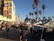 A busy outdoor marketplace with numerous people walking along a street lined with vendor stalls. Tall palm trees tower over the scene, adding a tropical atmosphere. A colorful mural is displayed on the side of a building, and a clear blue sky suggests a sunny day.