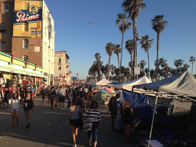 A busy outdoor marketplace with numerous people walking along a street lined with vendor stalls. Tall palm trees tower over the scene, adding a tropical atmosphere. A colorful mural is displayed on the side of a building, and a clear blue sky suggests a sunny day.
