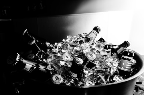 A collection of beer bottles immersed in ice within a large container. The scene is predominantly monochrome, providing a classic and timeless feel. The condensation and ice suggest a cold and refreshing atmosphere.