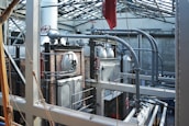 A complex industrial setting featuring large metal machinery, pipes, and valves within a sturdy brick and metal framework. The structure is under a glass roof that allows natural light to illuminate the area, highlighting the metallic texture of the equipment and the stark industrial architecture.