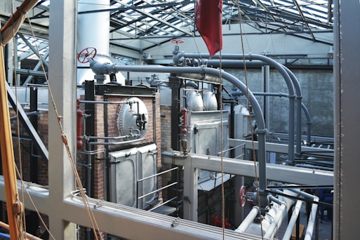A complex industrial setting featuring large metal machinery, pipes, and valves within a sturdy brick and metal framework. The structure is under a glass roof that allows natural light to illuminate the area, highlighting the metallic texture of the equipment and the stark industrial architecture.