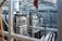 A complex industrial setting featuring large metal machinery, pipes, and valves within a sturdy brick and metal framework. The structure is under a glass roof that allows natural light to illuminate the area, highlighting the metallic texture of the equipment and the stark industrial architecture.