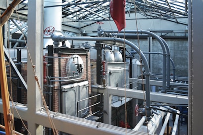 A complex industrial setting featuring large metal machinery, pipes, and valves within a sturdy brick and metal framework. The structure is under a glass roof that allows natural light to illuminate the area, highlighting the metallic texture of the equipment and the stark industrial architecture.