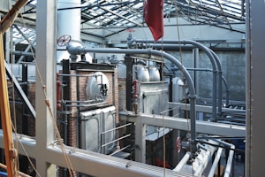 A complex industrial setting featuring large metal machinery, pipes, and valves within a sturdy brick and metal framework. The structure is under a glass roof that allows natural light to illuminate the area, highlighting the metallic texture of the equipment and the stark industrial architecture.