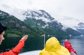 People wearing rain jackets point towards a scenic landscape of misty green mountains rising from a calm body of water. A small Norwegian flag is visible at the stern of a boat.