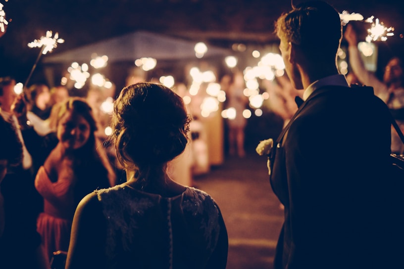 A couple stands together, with their backs facing the camera, surrounded by people holding sparklers. The scene is filled with guests celebrating in a warm and festive atmosphere likely a wedding reception or party. Soft lighting and sparkling lights create an intimate and joyful mood.
