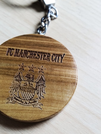 A wooden keychain featuring the engraved text 'FC Manchester City' along with an engraved emblem that includes stars and wings. The keychain is attached to a metallic key ring placed on a light wooden surface.