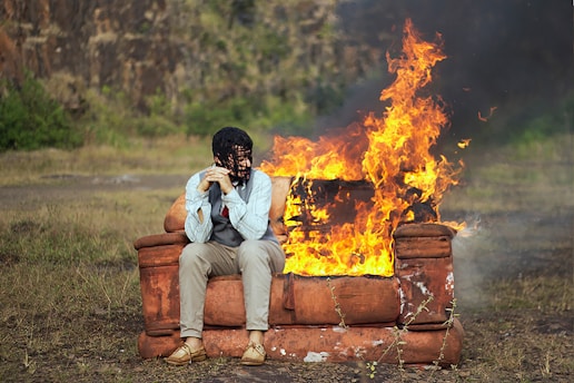 A person sits on an orange couch in an outdoor setting with a large fire engulfing part of the couch. The individual is hunched over with their head resting in their hands, surrounded by a grassy field and rocky background.