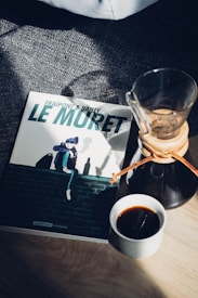 A graphic novel titled 'Le Muret' rests on a textured surface next to a cup of black coffee and a glass Chemex coffee maker. Sunlight casts shadows across the cover and surfaces, adding contrast and depth.