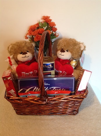 A wicker basket filled with an assortment of gifts including two plush teddy bears holding red heart-shaped pillows with 'I Love You' written on them. The basket also contains various chocolates such as Maltesers, Cadbury Milk Tray, and Kinder chocolates. A bouquet of orange roses sits behind the teddy bears.